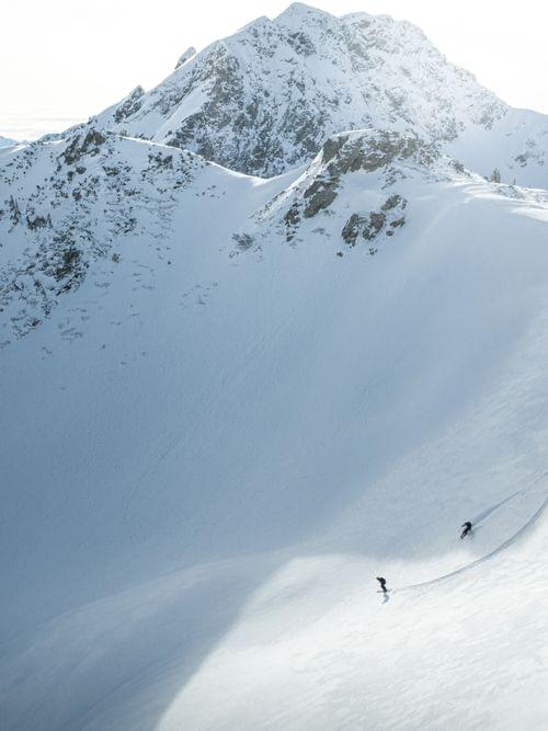 Zwei Skifahrer fahren eine verschneite alpine Piste mit Berggipfeln im Hintergrund hinab