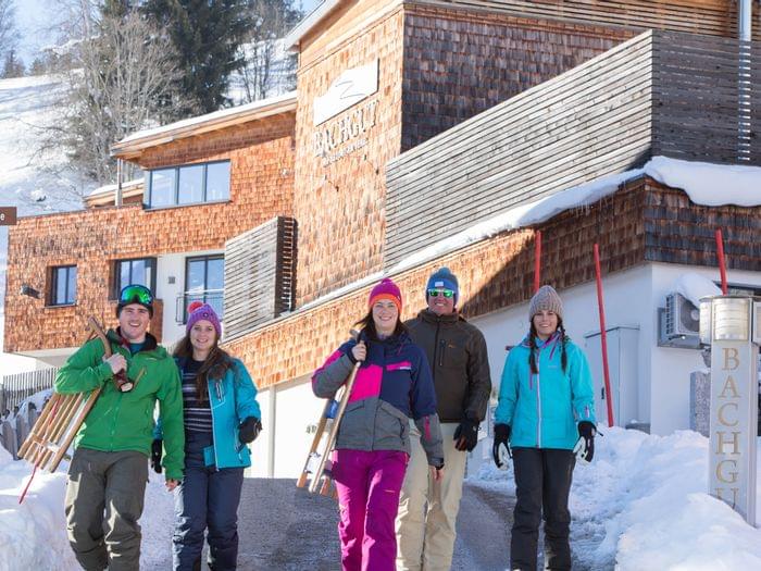 Group of smiling people in winter clothing walking with sleds near alpine lodge in snowy landscape