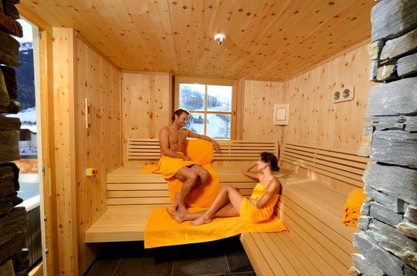 Paar entspannt in einer Holzsauna mit orangenen Handtüchern und Bergblick