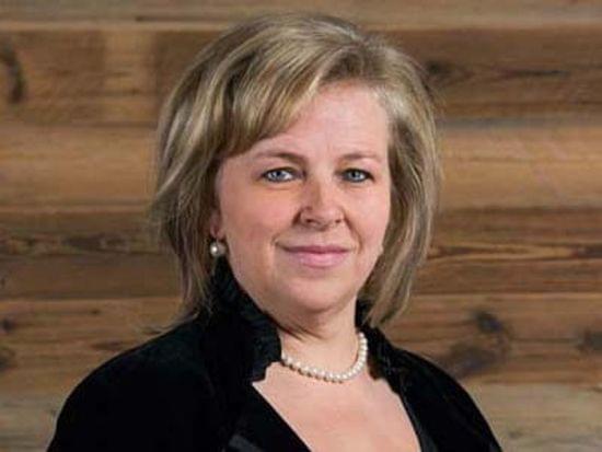 Smiling woman in formal black attire with pearl necklace standing against wooden wall