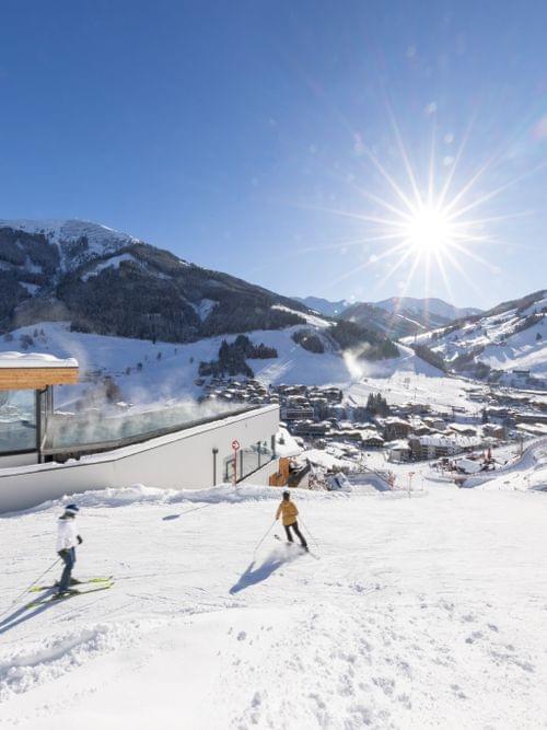 Skifahrer die neben dem Bachgut hinunter Fahren, im Blick das Badehaus mit Pool und das Ortszentrum von Hinterglemm 