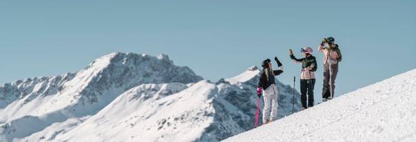 Drei Frauen in Skikleidung geben sich High-Five auf verschneiter Piste mit Berggipfeln im Hintergrund