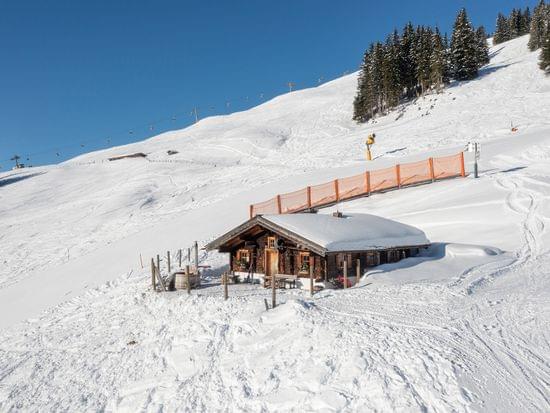 Holzhütte in den Alpen im Schnee auf sonniger Skipiste mit Skilift im Hintergrund