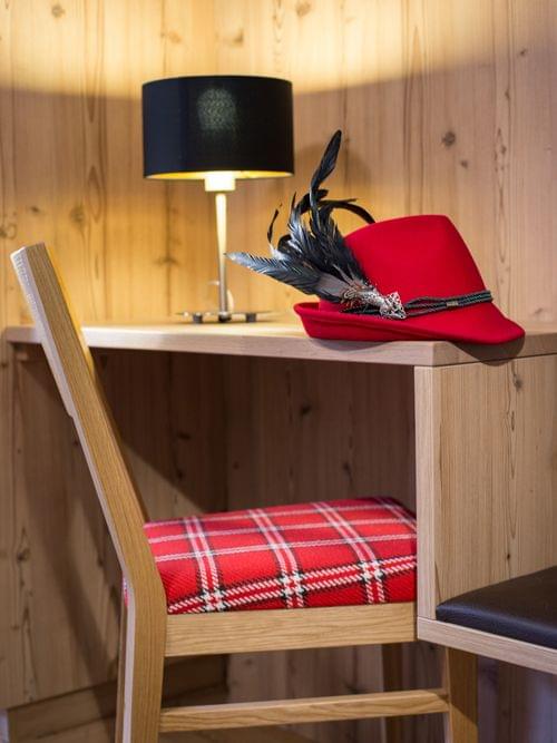 Roter traditioneller Alpenhut mit Federn auf Holzschreibtisch neben kariertem Stuhl