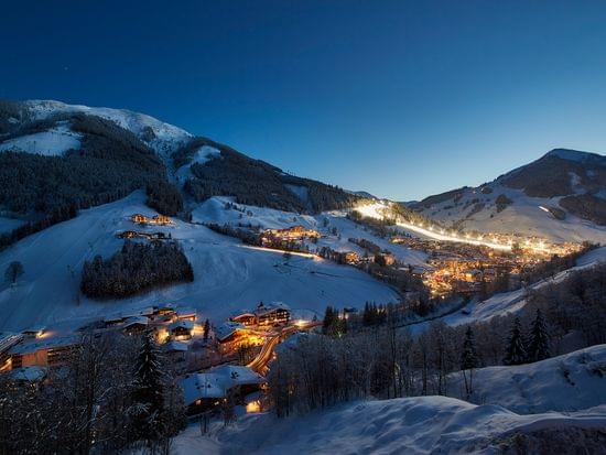 Snow-covered alpine village at dusk with illuminated ski slopes and mountain landscape