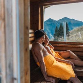 2 Personen in der Sauna, schauen durch das Panoramafenster auf die Bergwelt 