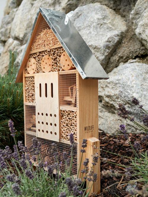 Wooden insect hotel with metal roof in garden near stone wall and lavender plants