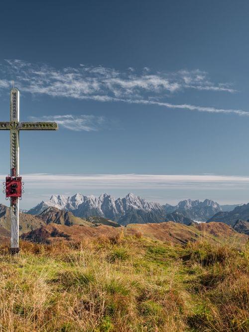 Gipfelkreuz auf grasbewachsenem Berggipfel mit Panoramablick auf verschneite Alpen unter blauem Himmel