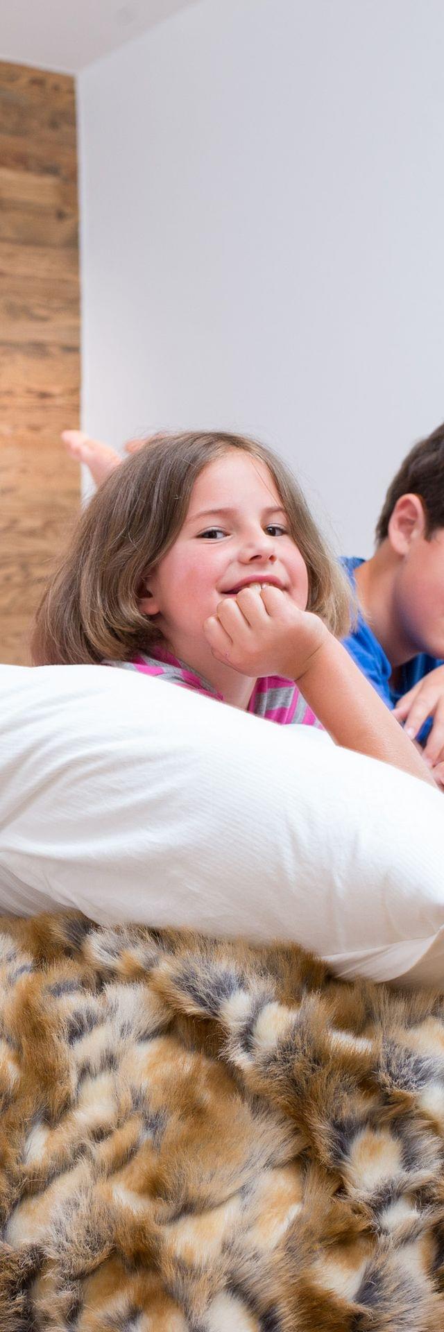 Drei lachende Kinder liegen auf einem gemütlichen Bett mit flauschiger Felldecke im rustikalen Schlafzimmer