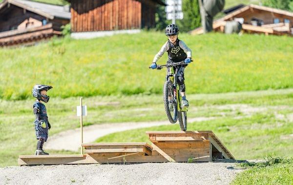 Junge springt mit Mountainbike über Holzrampe, ein anderes Kind schaut in Schutzausrüstung zu