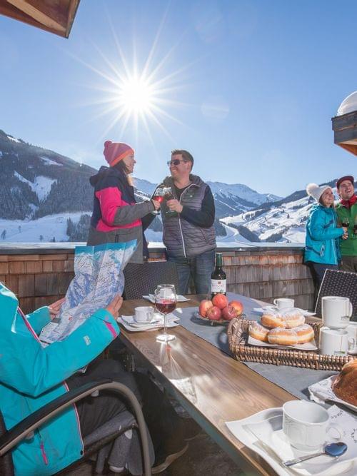 Group enjoying drinks and pastries on a sunny ski chalet terrace with alpine mountain views