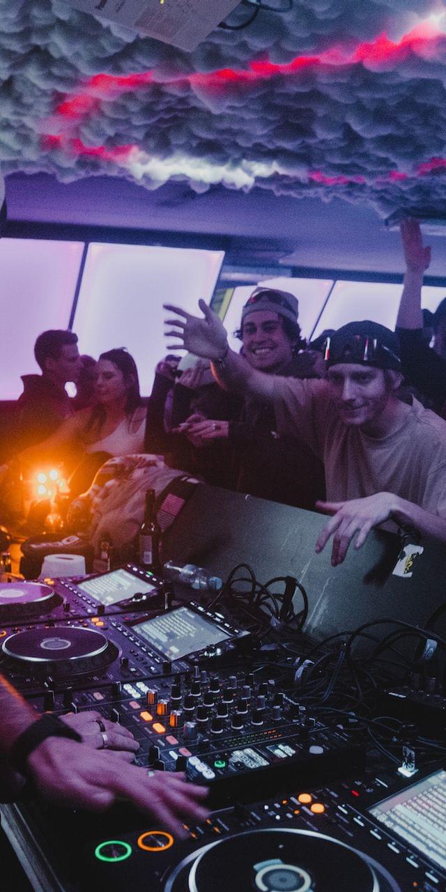 DJ vor seinem Pult, davor Gäste die Tanzen - Club Stimmung, beleuchtete Decke 