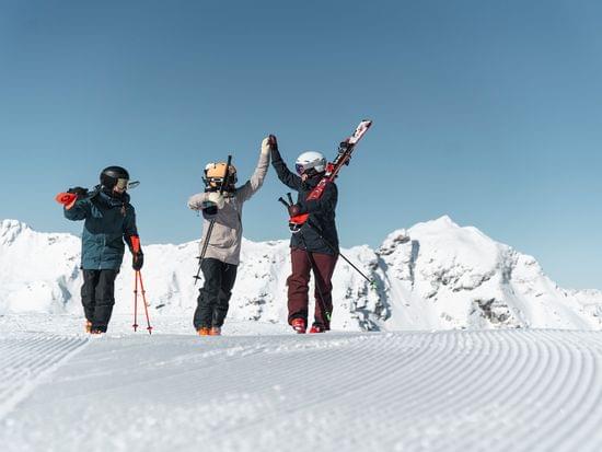 Drei Skifahrer feiern auf einer verschneiten Piste mit Skiern und Stöcken unter blauem Himmel