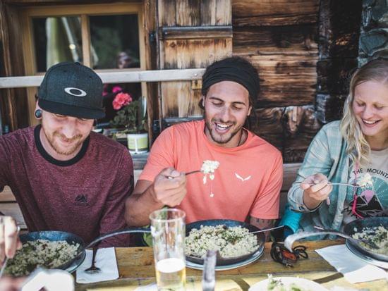 2 Männer und 1 Frau sitzen am Tisch vor einer Almhütte beim Mittagessen 