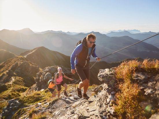 Gruppe von Wanderern steigt im Sonnenaufgang einen steilen Bergpfad mit Seilsicherung hinauf