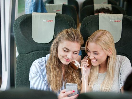 Two young women sharing headphones and watching a smartphone on an ÖBB Railjet train