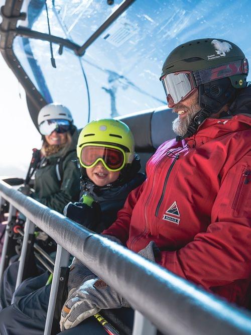 Familie in Skibekleidung fährt bei Sonnenschein im Skigebiet mit dem Sessellift