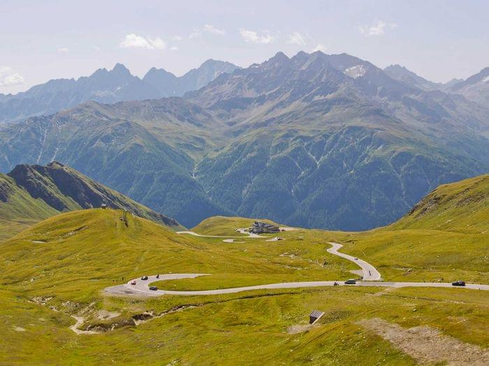 Winding mountain road through green alpine landscape with distant peaks in the background