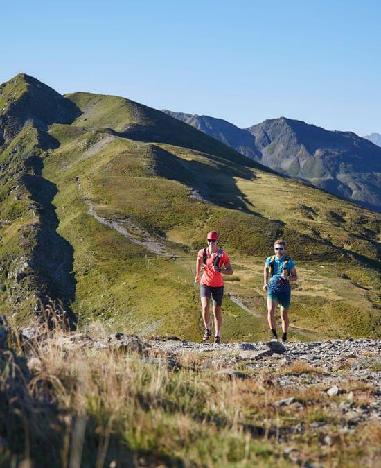 Trail runners on a mountain ridge path with alpine peaks in the background