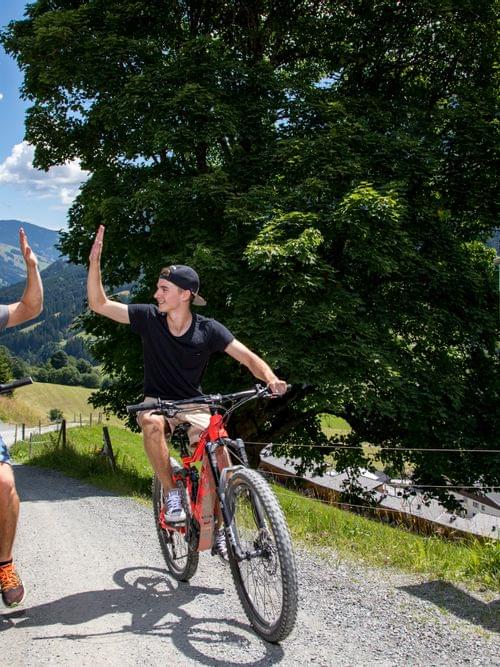 Two young men high-five while mountain biking on a scenic alpine trail near chalets
