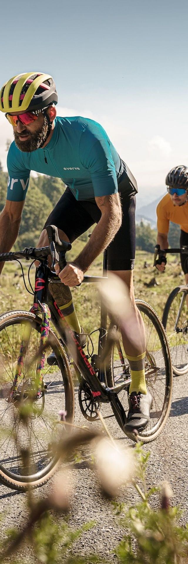 3 Gravel Biker die eine Schotterstraße fahren, Ringsum Berge und Almwiesen