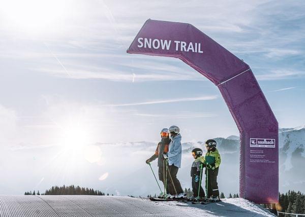 2 Erwachsene mit 2 Kindern beim Skifahren, Sie stehen vor einem Schild mit dem Namen Snow Trail 