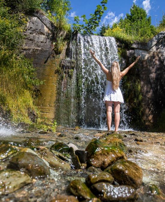 Frau im weißen Kleid steht vor Wasserfall im grünen Wald an einem sonnigen Tag