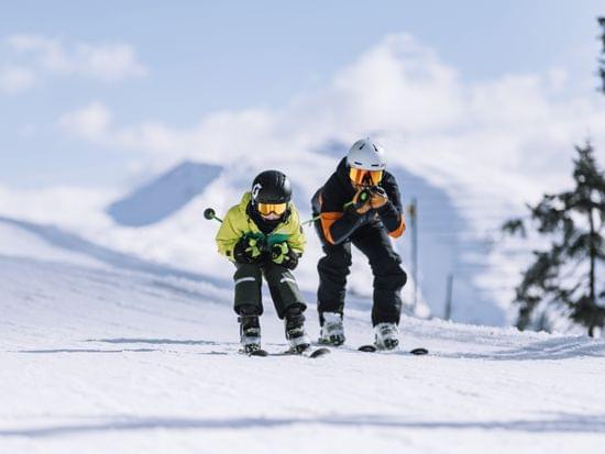 Two children skiing downhill in racing posture on a sunny snowy mountain slope