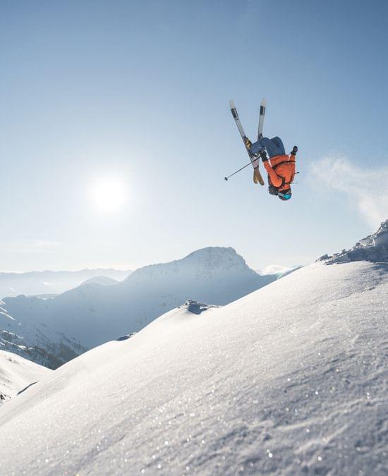 Freestyle-Skifahrer macht Rückwärtssalto über verschneitem Berghang bei blauem Himmel