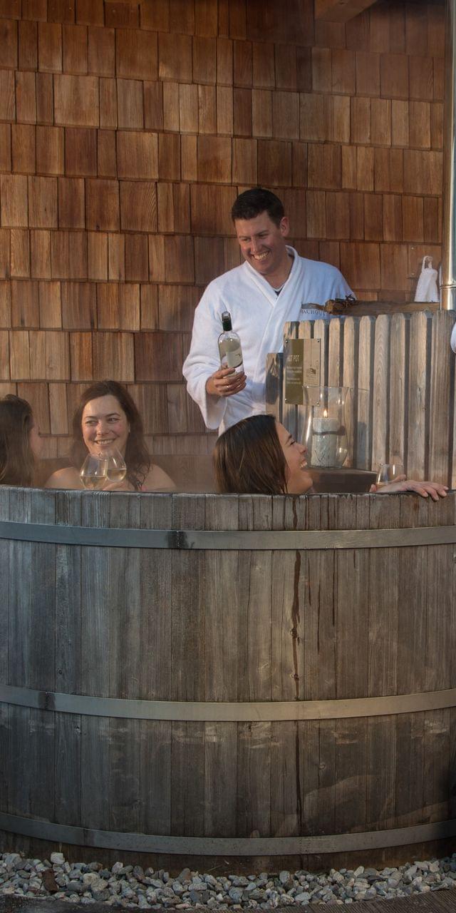 Freunde entspannen im hölzernen Außen-Whirlpool mit Wein und Bademänteln