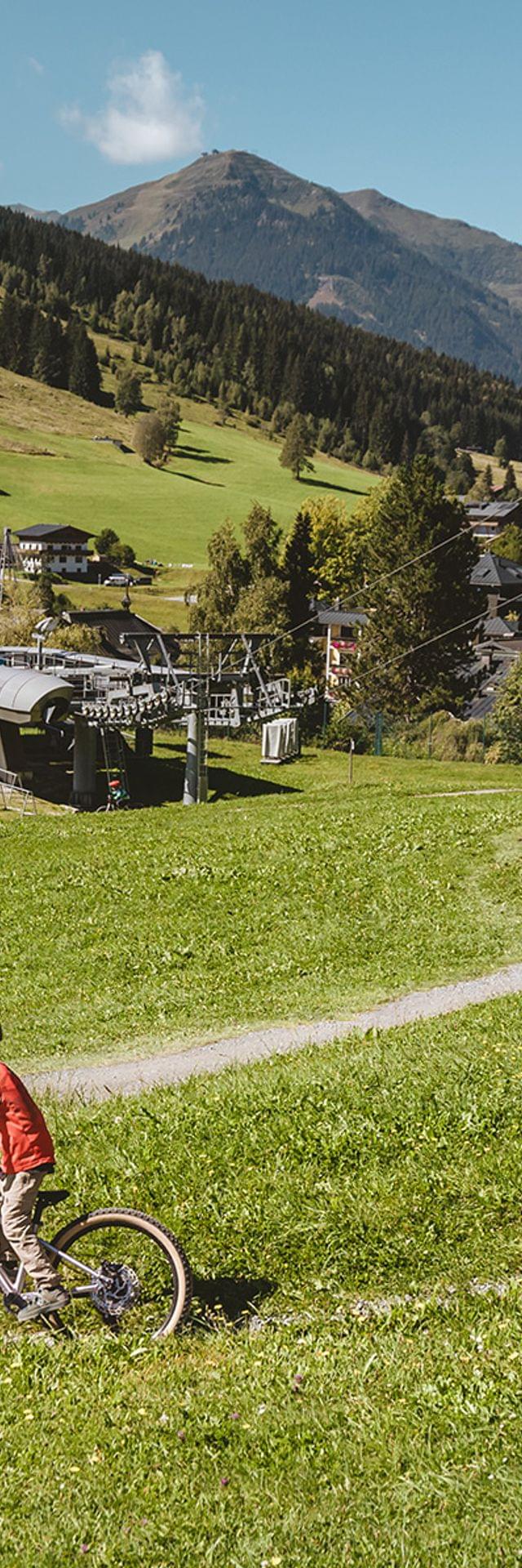 Familie beim Mountainbiken auf einem Trail im Alpendorf mit Seilbahn und Bergen im Hintergrund