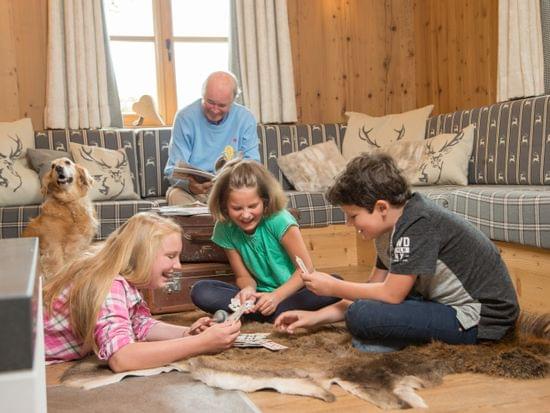 Kinder spielen Karten auf einem Fellteppich im gemütlichen Wohnzimmer mit Hund und älterem Mann beim Lesen