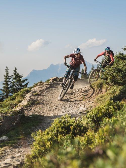 Two mountain bikers riding downhill on a rugged trail surrounded by alpine scenery