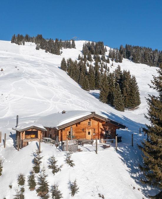 Holzhütte in den Alpen umgeben von verschneiten Hängen und Tannen unter klarem blauen Himmel
