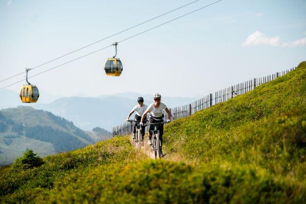 2 Personen mit den Mountainbikes am Berg auf einem Trail im Hintergrund fährt die Gondelbahn 