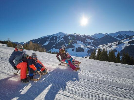 Familien rodeln eine verschneite Piste hinab bei Sonnenschein im Skigebiet