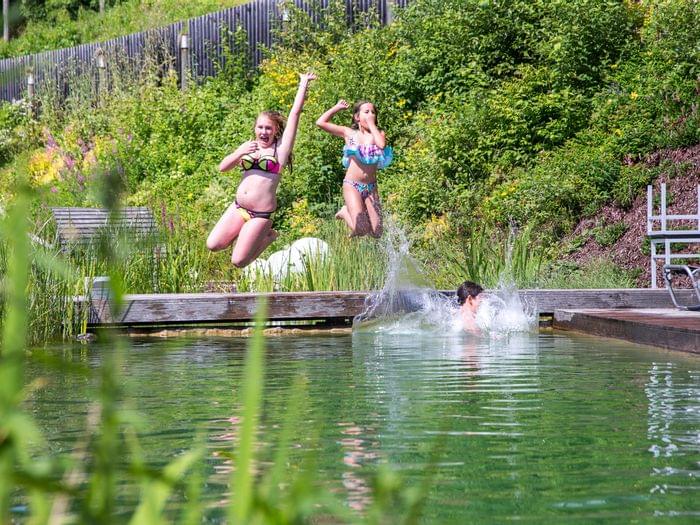 Children jumping into a natural swimming pond surrounded by greenery on a sunny day