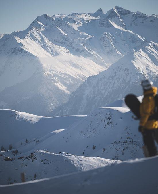 Snowboarder in yellow jacket overlooking snowy alpine mountain range on sunny day