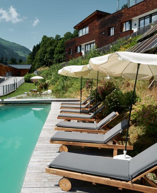 Naturschwimmteich mit Liegen und Sonnenschirmen und dahinter die Bachgut Chalets zu sehen, Sommer und Sonnenschein 