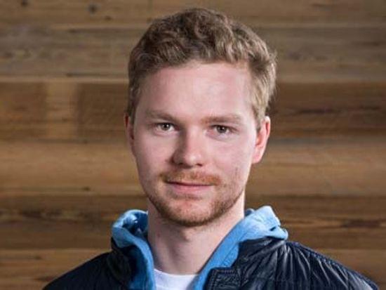 Mann in blauem Hoodie und schwarzer Jacke vor hölzerner Wand