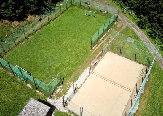 Luftaufnahme eines eingezäunten Fußballfelds aus Gras und eines Sand-Volleyballfelds im Waldgebiet