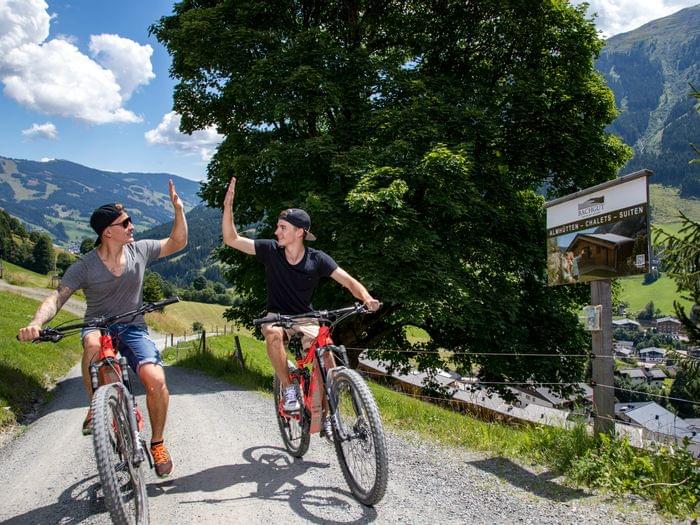 Zwei junge Männer geben sich beim Mountainbiken auf einem alpinen Weg ein High-Five nahe Chalets