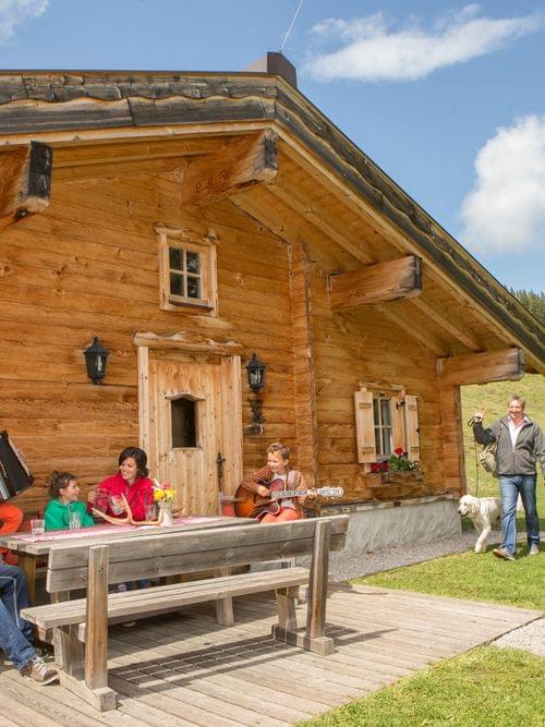 Family enjoying music and drinks outside a rustic wooden alpine cabin in summer