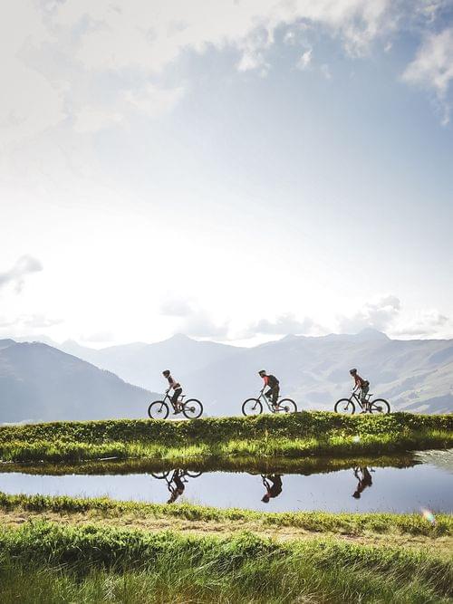 Drei Mountainbiker auf einem Pfad neben einem spiegelnden Teich vor Alpenkulisse
