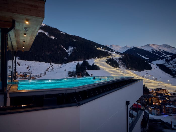 The Bachgut bathhouse infinity pool overlooking the floodlit U-Bahn ski slope, at dusk in a snow-covered winter setting.