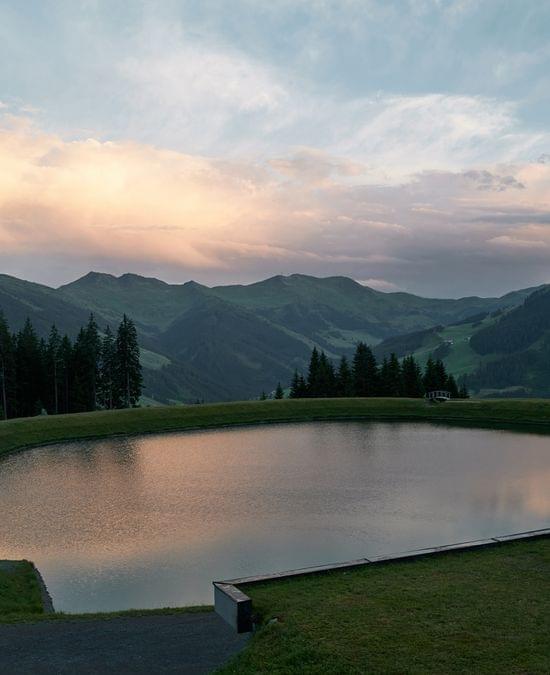 Bergsee mit Spiegelung des Sonnenuntergangs vor Wald und Alpenhügeln