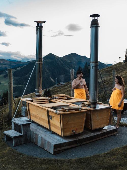 Two people in front of the outdoor bathtubs, with a twilight sky and alpine meadows in the background.