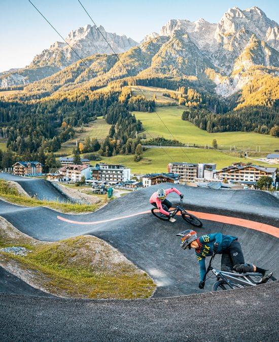 Two mountain bikers riding a pump track in alpine village with scenic mountain backdrop