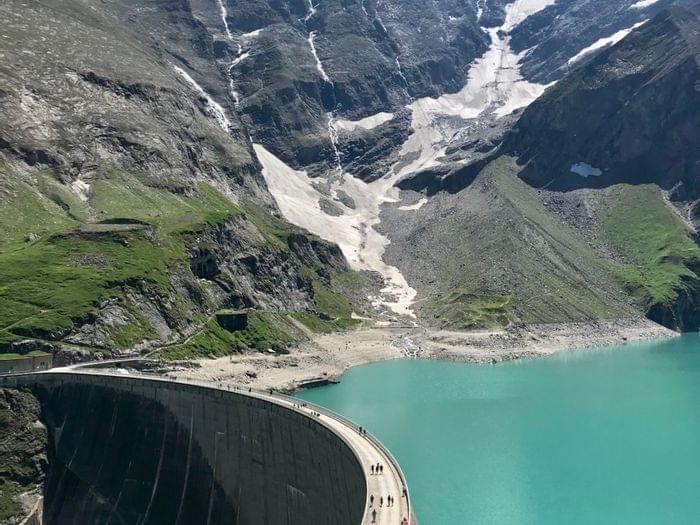Gebogener Hochgebirgsstausee mit türkisfarbenem Wasser und schneebedeckten Gipfeln im Sommer