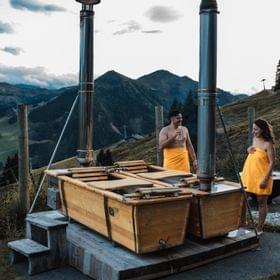 2 Personen vor den Außenbadewannen im Freien, dahinter dämmernder Himmel und Almwiesen 